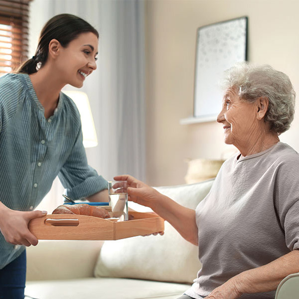 A young woman smiles as she hands a breakfast tray with coffee and croissants to an elderly woman sitting on a couch in a cozy living room.