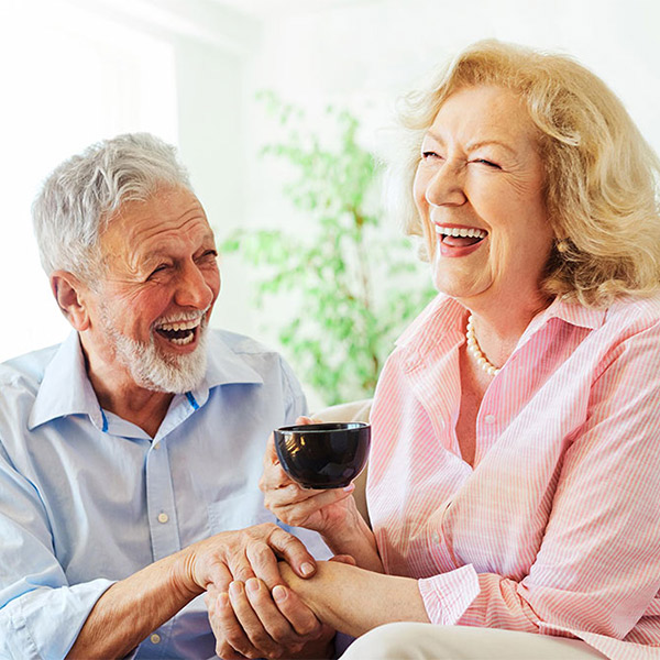 An elderly man and woman sit together, smiling and laughing. The woman holds a cup while the man gently holds her hand. They appear happy and relaxed in a bright, indoor setting with green plants in the background.