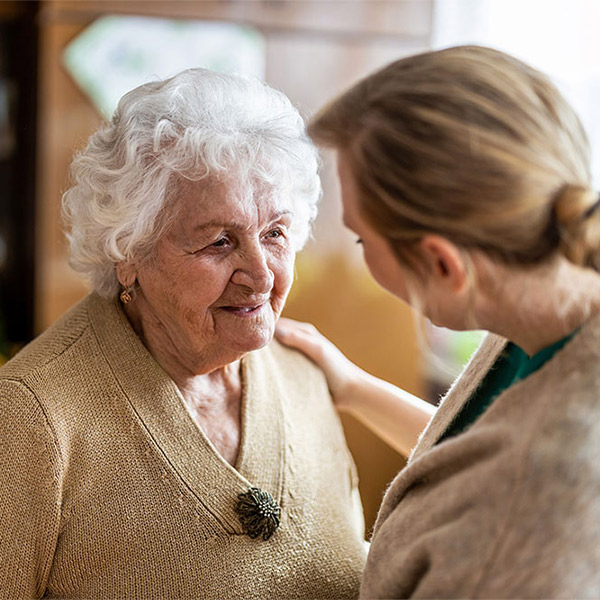 An elderly woman with white hair smiles warmly while a younger woman gently touches her shoulder, offering comfort and support in a cozy indoor setting.
