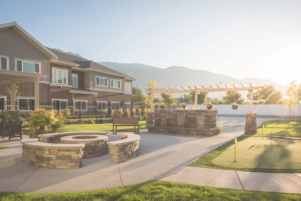 A backyard with a stone fire pit, curved seating, a pergola with hanging plants, a putting green, and a large house in the background, all illuminated by bright sunlight.
