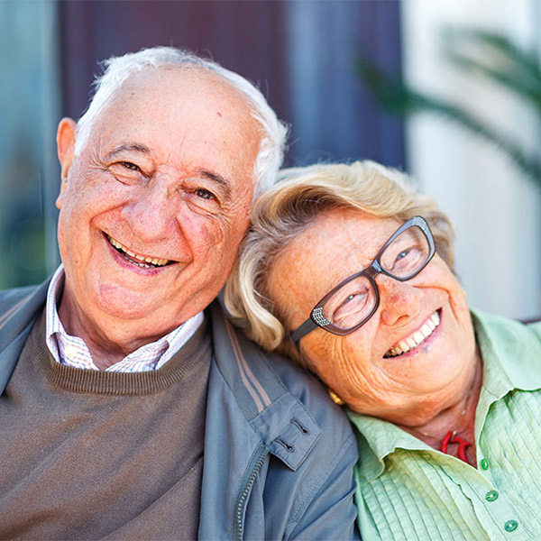 An elderly couple sits closely together, smiling warmly. The man wears a gray jacket and striped shirt, while the woman, in glasses and a green blouse, leans her head on his shoulder. They appear happy and relaxed.