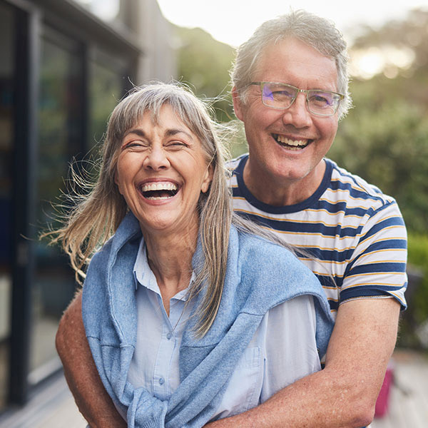An older couple stands outside, smiling and laughing. The man is giving the woman a playful hug from behind. Both look happy and relaxed, enjoying a sunny day.