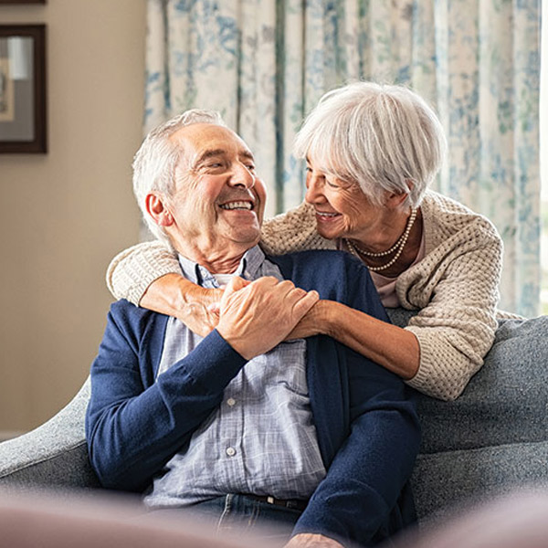 An elderly couple sits together on a couch, smiling and embracing warmly. The woman wraps her arms around the man from behind, and both look happy and content in a cozy, well-lit living room.