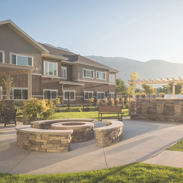 A modern two-story house with large windows, a fenced yard, and a landscaped outdoor area featuring curved stone benches and a fire pit, with mountains visible in the background.